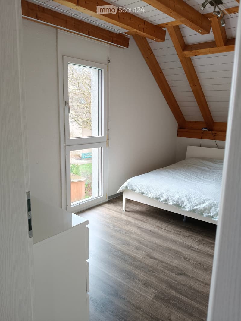 Cozy bedroom with wooden slanted ceiling beams, a white bed with a light blue duvet, and a white cabinet. The room features wooden flooring and large windows with a view of the garden.