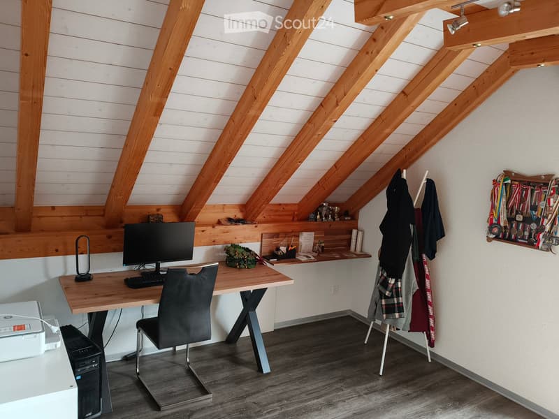 A modern, well-lit attic workspace with wooden beams, a sleek desk setup, and a cozy chair. Shelves hold various items, including medals and a small plant.