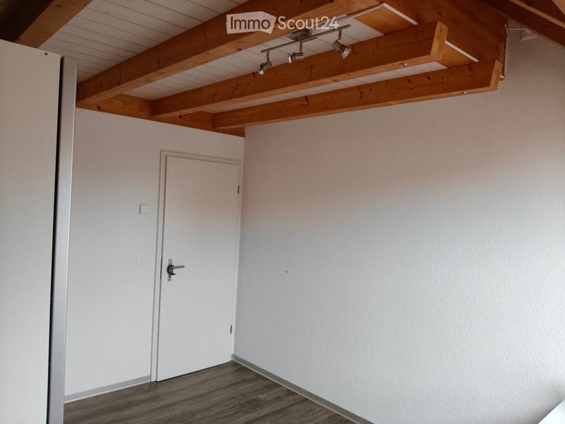 Empty room with exposed wooden beams on the ceiling, white walls, and a wooden floor. A closed door is visible on the left side.