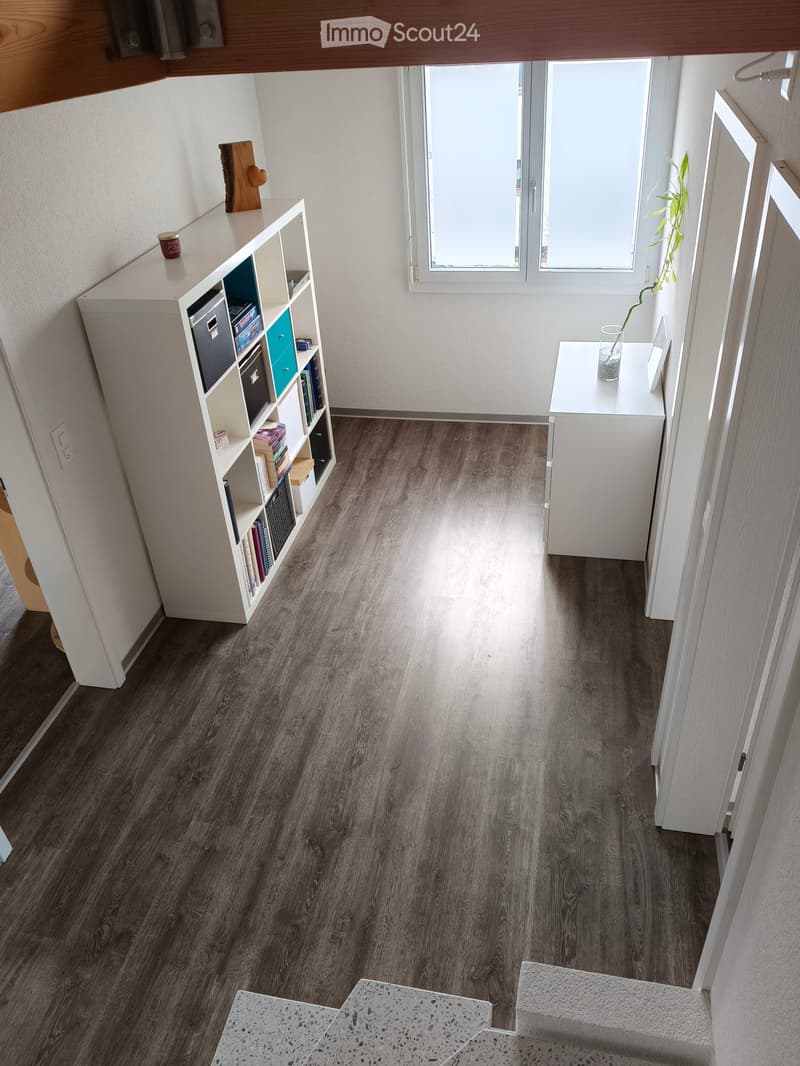 A modern hallway with dark wood flooring, white walls, and a staircase leading upwards. Features include a built-in shelving unit with books and storage boxes, a small table with a vase, and a window letting in natural light.