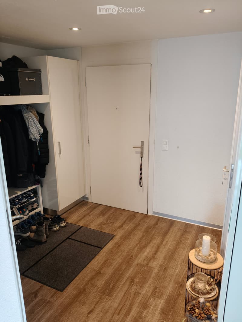 The image shows a hallway with wooden flooring, a small round stool with a candle, a shoe rack with shoes, and a closet with clothes and storage boxes.