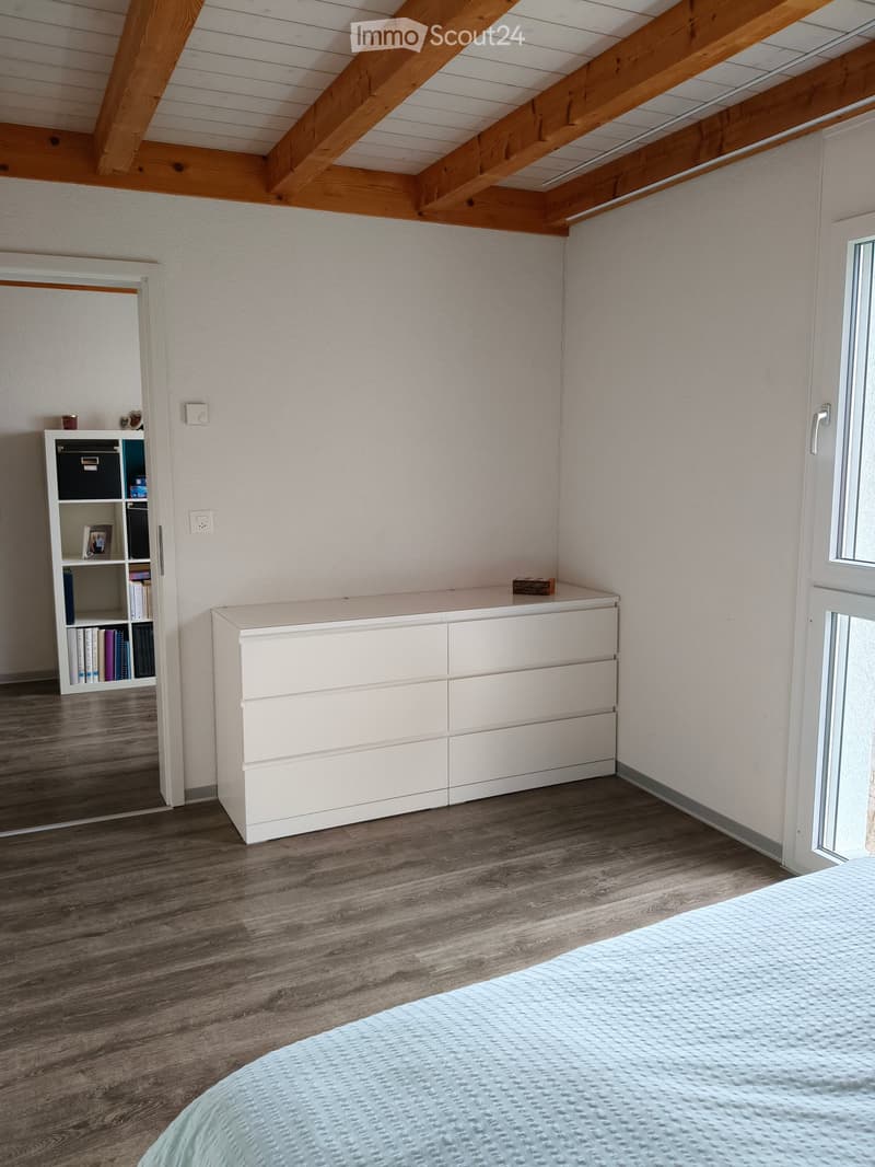 Bedroom with a light blue bedspread, white cabinet, bookshelf, and exposed wooden beams on the ceiling.