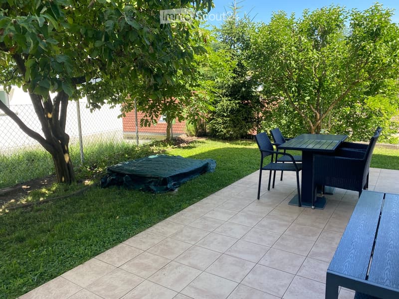 Garden area with tiled patio, outdoor table and chairs, bench, covered object, and trees.