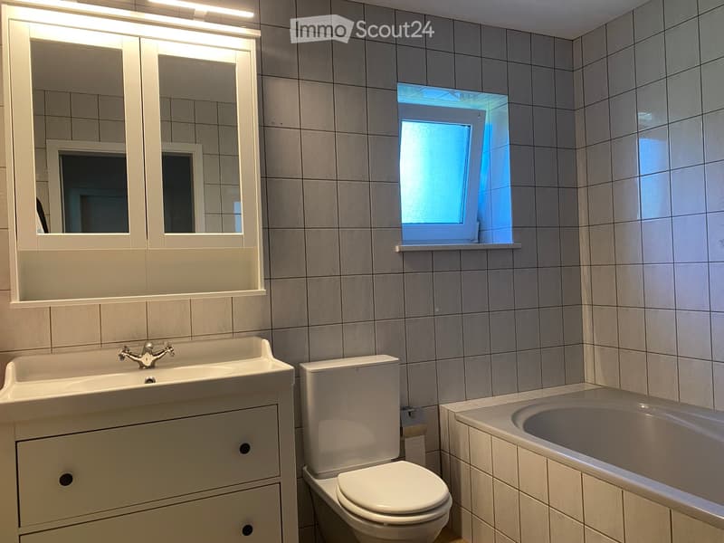 Modern bathroom with a double sink, toilet, bathtub, and large mirror with built-in storage. The room features tiled walls and a window letting in natural light.