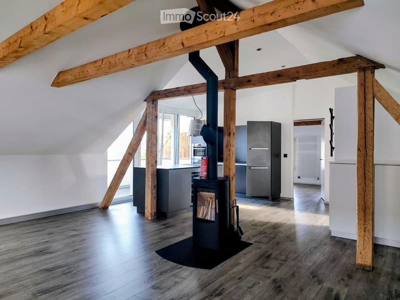 Open-plan attic space with exposed wooden beams, wooden floor, fireplace, modern kitchen area with refrigerator and oven, and large windows letting in natural light.