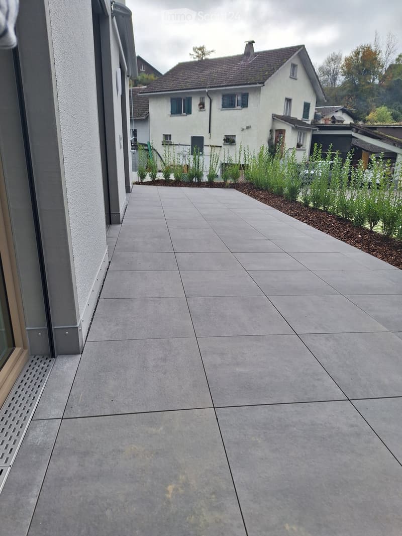 Gray tiled outdoor walkway with glass doors and gray plants in the background.
