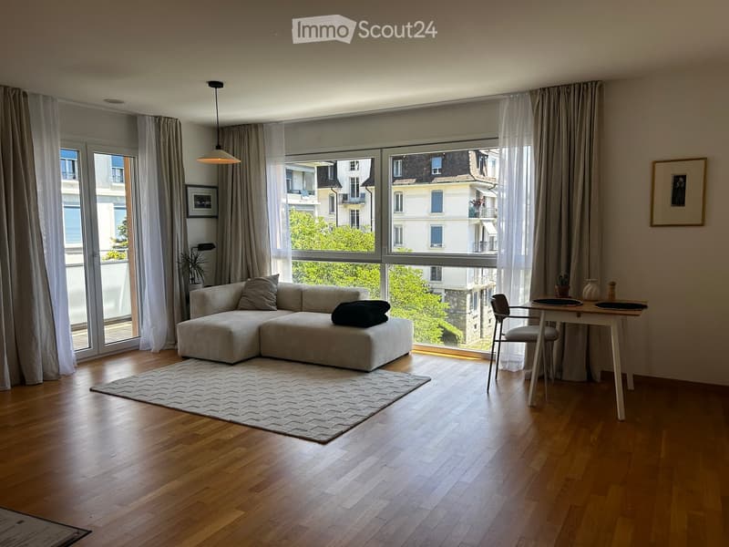 Modern living room with large windows, wooden flooring, a sectional sofa, a coffee table, a dining table with chairs, and decorative items.