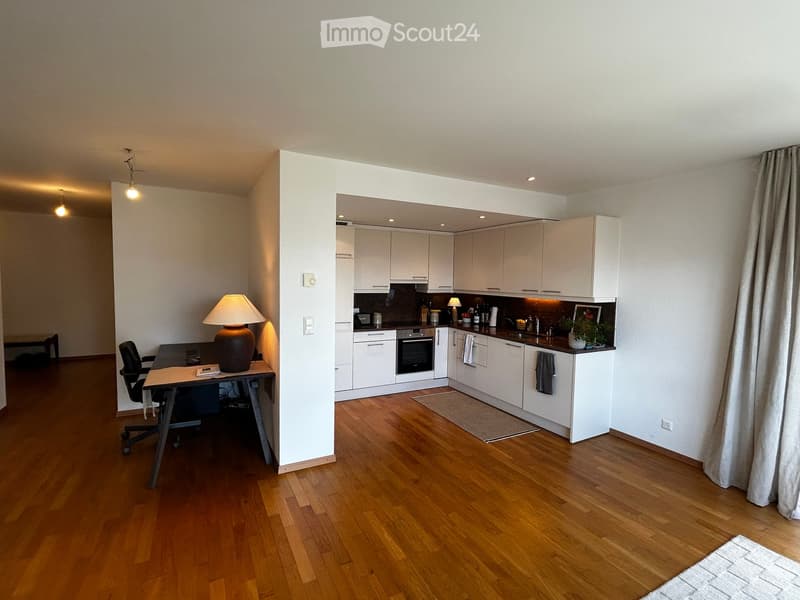 Open-plan living and kitchen area with wooden flooring, white cabinets, a desk with a chair, a lamp, and a small plant on the kitchen counter.
