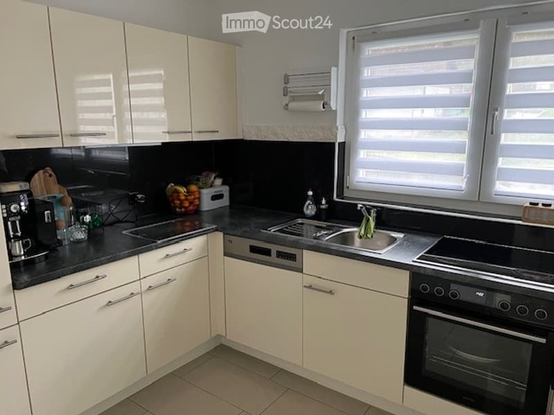 Modern kitchen with white cabinets, black countertops, and stainless steel appliances including a coffee maker, fridge, oven, sink, and dishwasher. The space features large windows with blinds and a bowl of fruit on the counter.