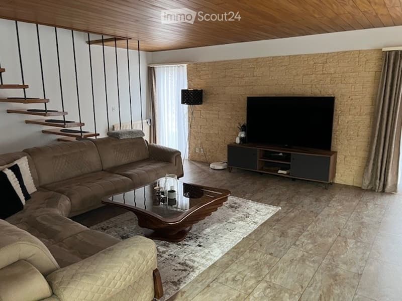 Modern living room with a beige sectional sofa, a glass coffee table, a TV stand, a floor lamp, and a curtained window. The room features a textured brick accent wall and a wooden ceiling.