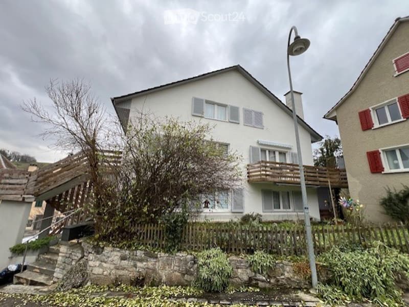 Two-story house with white exterior, multiple windows, raised ground floor, and a wooden balcony on the upper level.