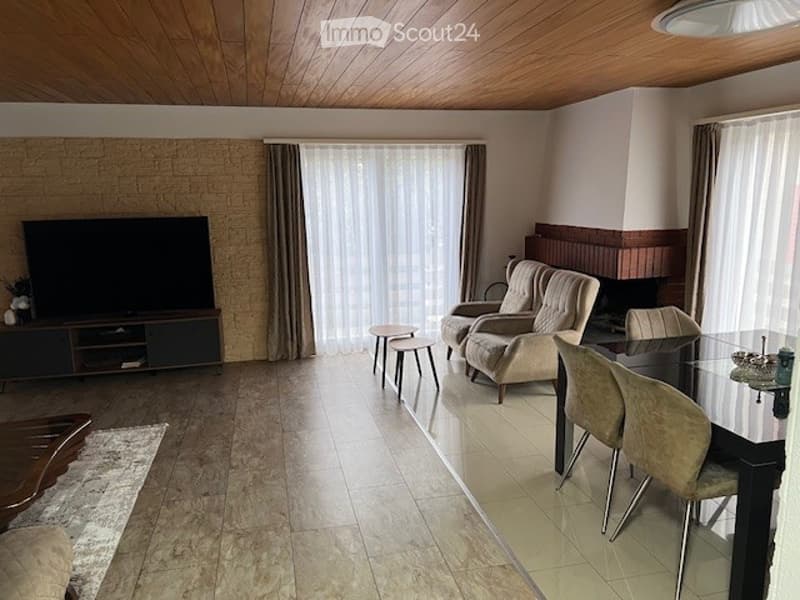 Spacious living room with wooden ceiling, fireplace, large windows with curtains, sofa, chairs, coffee table, TV, and dining area.
