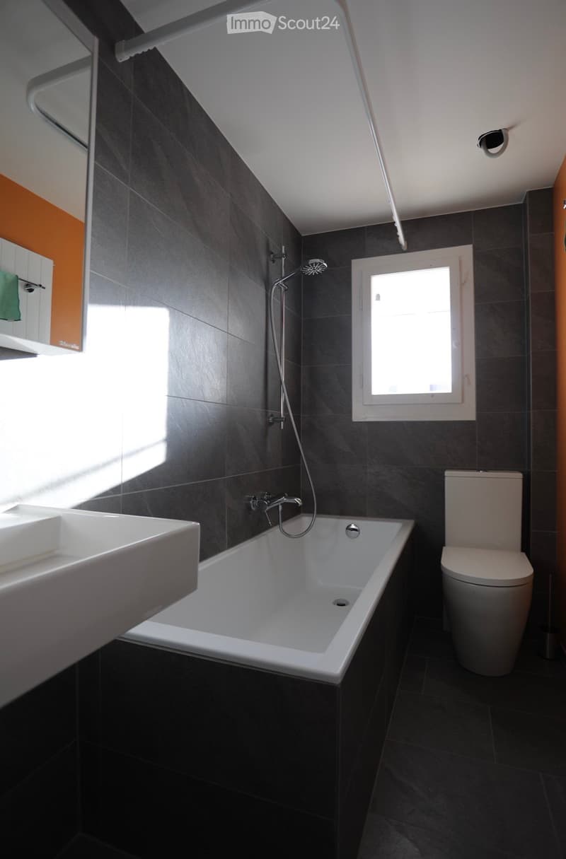 Modern bathroom with dark grey tiles, a white bathtub, a toilet, a sink, and a showerhead. A window provides natural light.