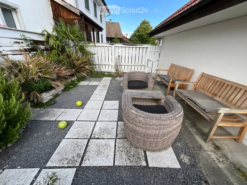 A small outdoor seating area with wicker chairs, a wooden bench, and a table, surrounded by plants and a white fence.