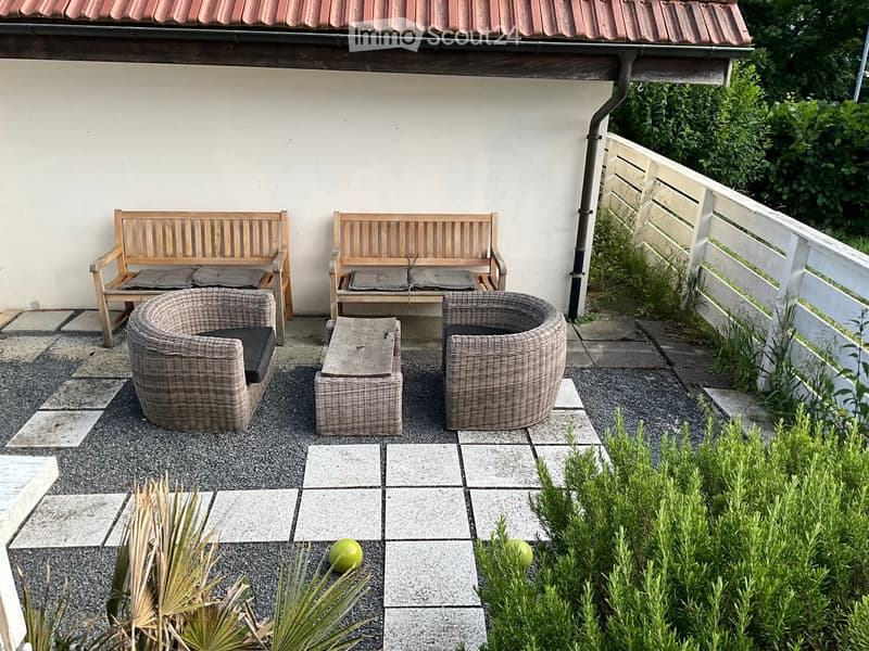 A small outdoor seating area with two wooden benches, two wicker chairs, and a small wooden table, surrounded by plants and a paved patio.
