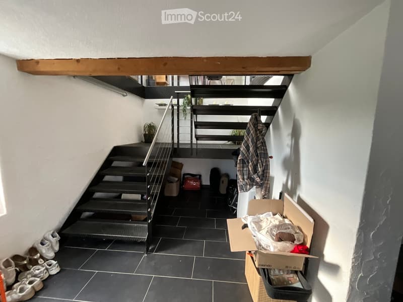 A narrow hallway with a staircase leading upwards, featuring black tiled flooring, white walls, and a metal railing. Several pairs of shoes are lined up against the wall, and a jacket is draped over a railing. Cardboard boxes and other items are stacked n