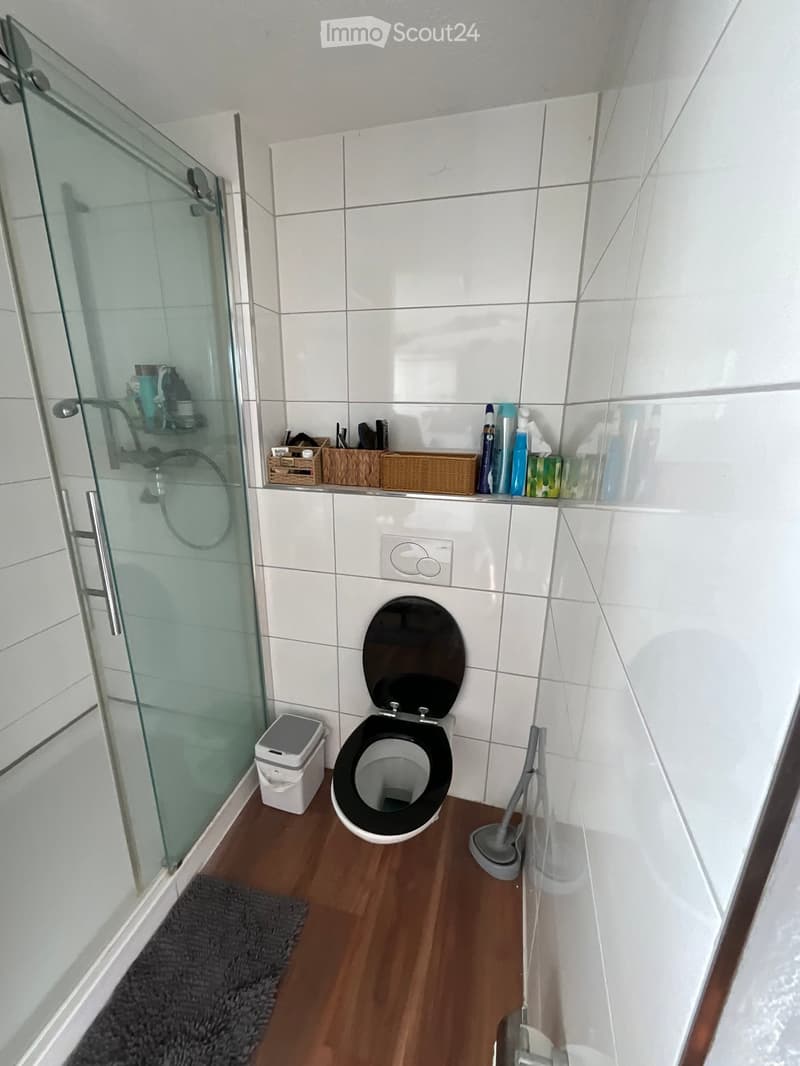 Modern bathroom with white tiled walls, a toilet, a glass-enclosed shower, a trash bin, and cleaning supplies on a shelf.