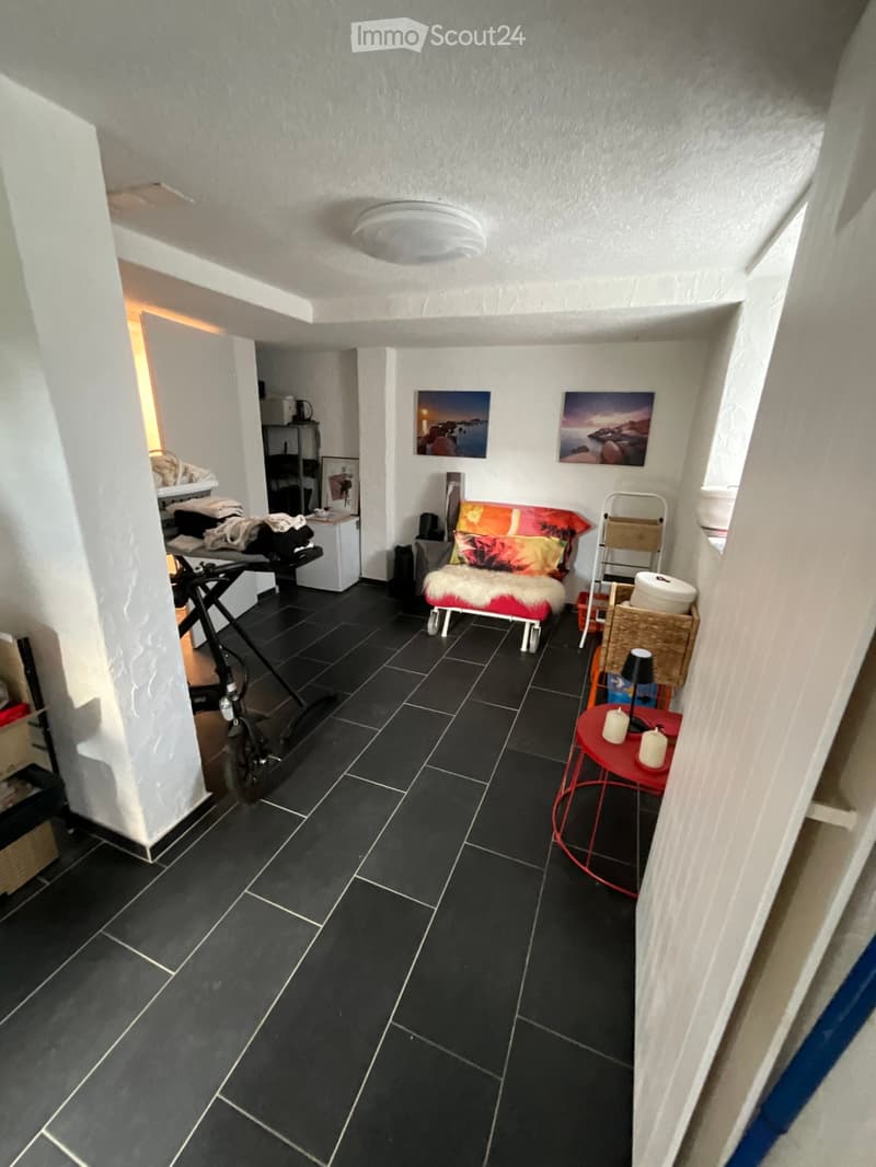 A small, modern hallway with black tiled flooring, white walls, and a ceiling light. Features a red metal table with candles, a colorful futon, a small shelf with a mirror, and a bicycle in the background.