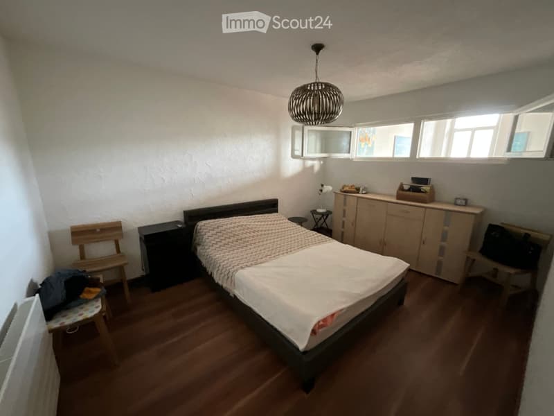 Cozy bedroom with wooden flooring, a double bed with a patterned duvet, a wooden chair, a small cabinet, a mirror, and a hanging lamp.