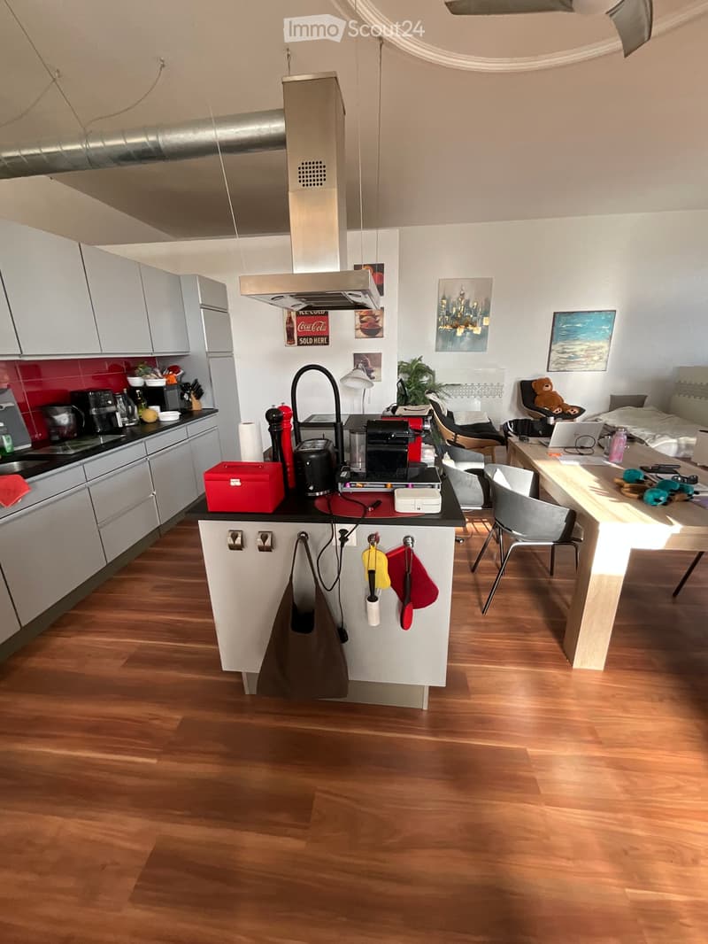Open-plan kitchen and living area with wooden flooring, white cabinets, a central island, and various appliances. A bed and desk are visible in the adjacent living space.