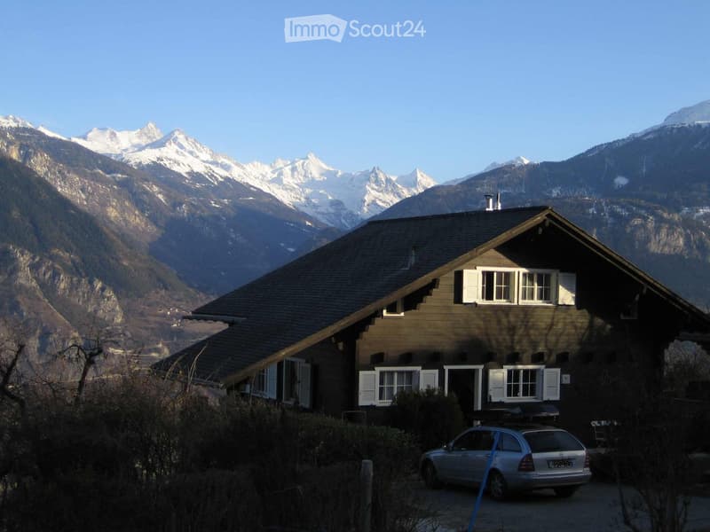 A two-story wooden house with white shutters and a dark roof, situated in a mountainous area with snow-capped peaks in the background. A silver car is parked in front of the house.