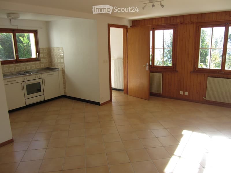 A partially furnished room with tiled flooring, wooden walls, and a kitchen area featuring an oven and cabinets. The room has large windows and a door leading to another space.