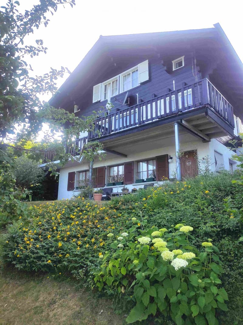 Two-story house with a balcony, surrounded by lush greenery and flowers.