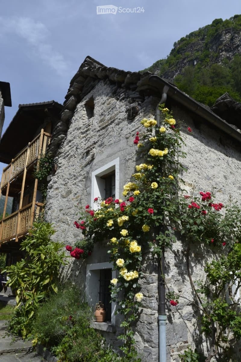 A rustic stone house with a wooden balcony, surrounded by vibrant yellow and red flowers, with a mountainous backdrop.