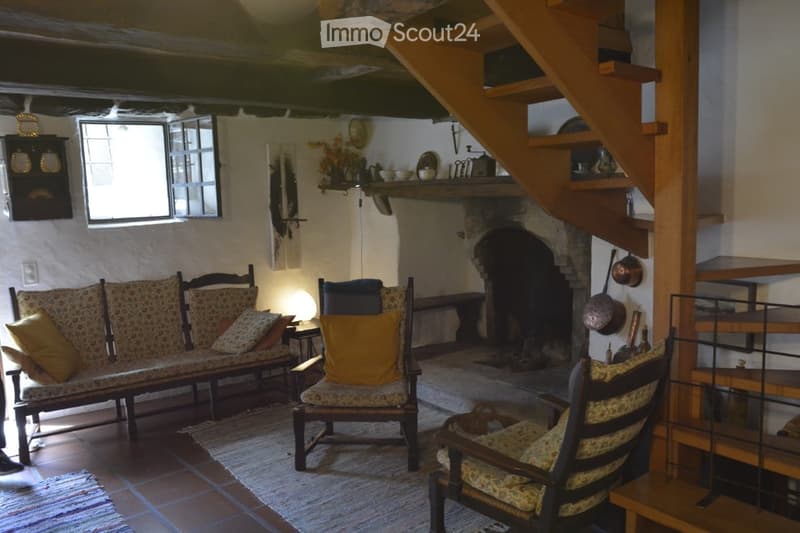 Cozy living room with exposed wooden beams, a stone fireplace, a patterned sofa, wooden chairs, and a small table. Shelves hold decorative items and kitchenware.