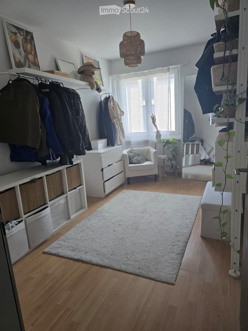A well-lit room with wooden flooring, a white chair, a storage unit with baskets, a coat rack, and a small plant near the window.