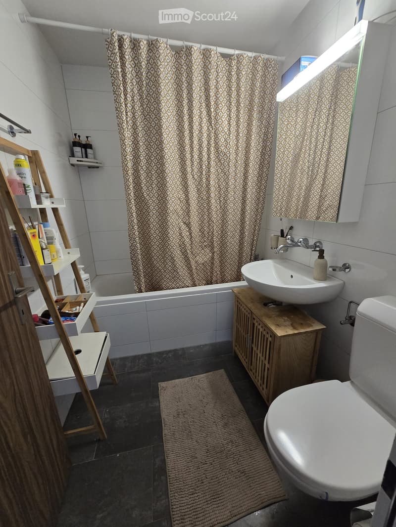 Modern bathroom with a white sink, toilet, bathtub, and a wooden shelf holding toiletries. A patterned shower curtain is visible, along with a bathroom rug on the floor.
