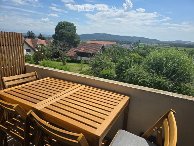 Balcony with wooden table and chairs, overlooking a scenic countryside with houses and trees.