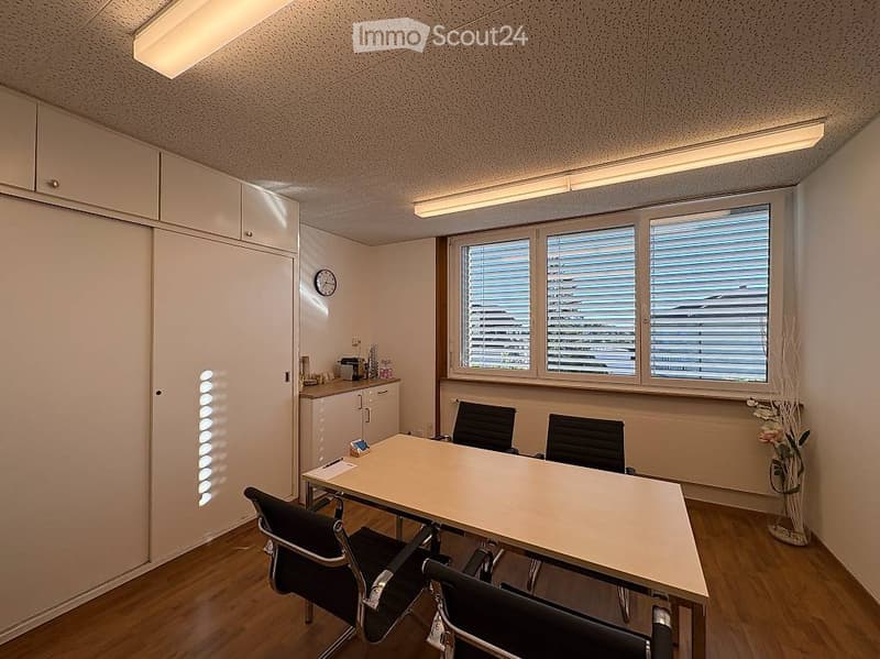 Office space with a large wooden table, four black chairs, white cabinets, a clock on the wall, and a vase with flowers near the window.
