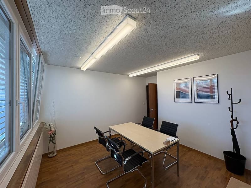 Bright, modern office space with wooden flooring, large windows, a table surrounded by chairs, and framed pictures on the wall.