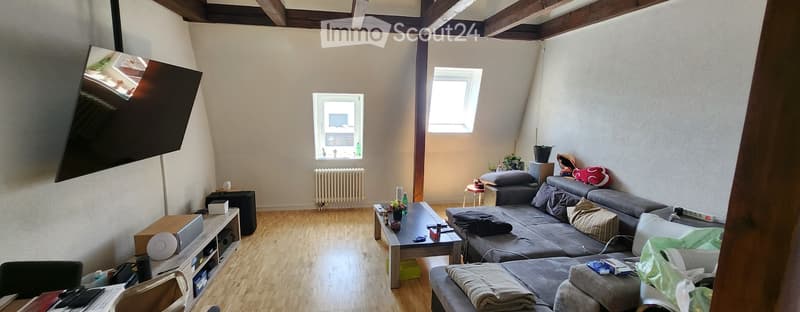 Living room with wooden flooring, exposed wooden beams on the ceiling, large windows, grey sofa, coffee table, TV stand, radiator, and small stool.