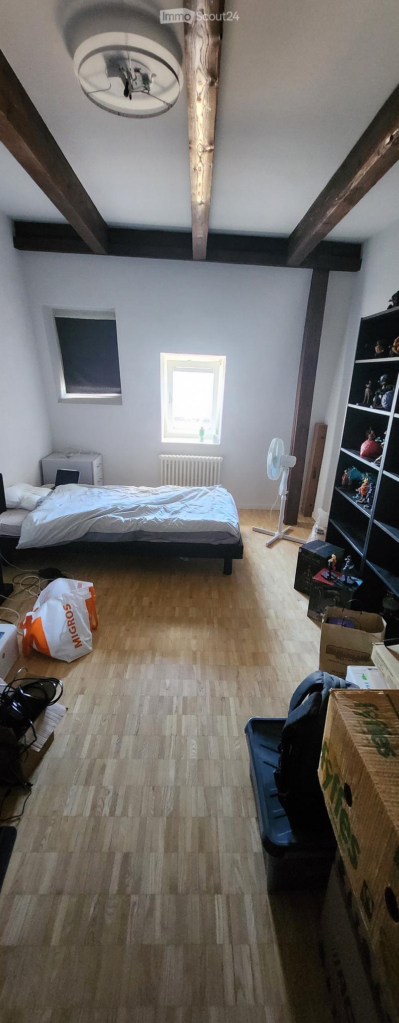A small, well-lit bedroom with wooden flooring, a bed, a fan, a shelf, and a laptop on a table.