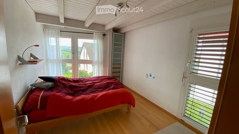 Bright bedroom with wooden flooring, a red duvet-covered bed, two pillows, a nightstand with a lamp, and large windows with white curtains. The room has white walls and a slanted wooden ceiling.