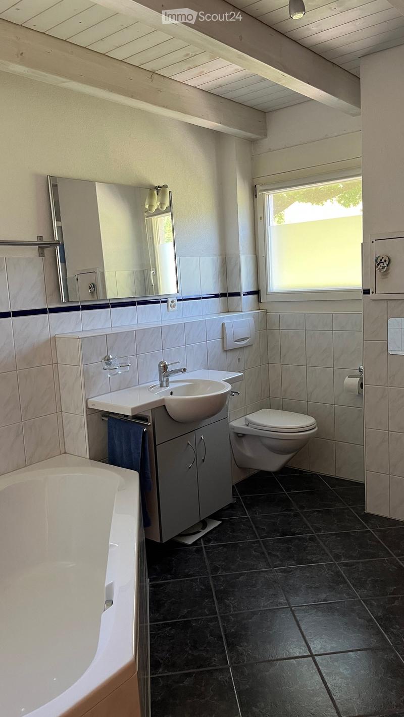 Modern bathroom with a white sink, toilet, bathtub, and mirror. Features dark tiled flooring and light-colored walls with blue accents.