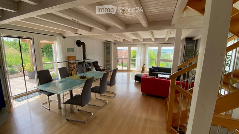 Bright, open-plan living and dining area with wooden floors, large windows, a red sofa, a glass dining table with chairs, and a fireplace.