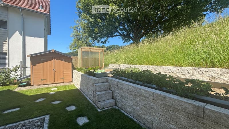 Well-maintained garden with a raised stone planter, wooden shed, and lush greenery, including tall grasses and trees, under a clear blue sky.