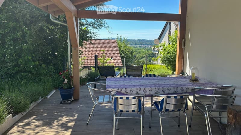 Wooden terrace with a table and chairs, overlooking a scenic landscape with trees and hills.