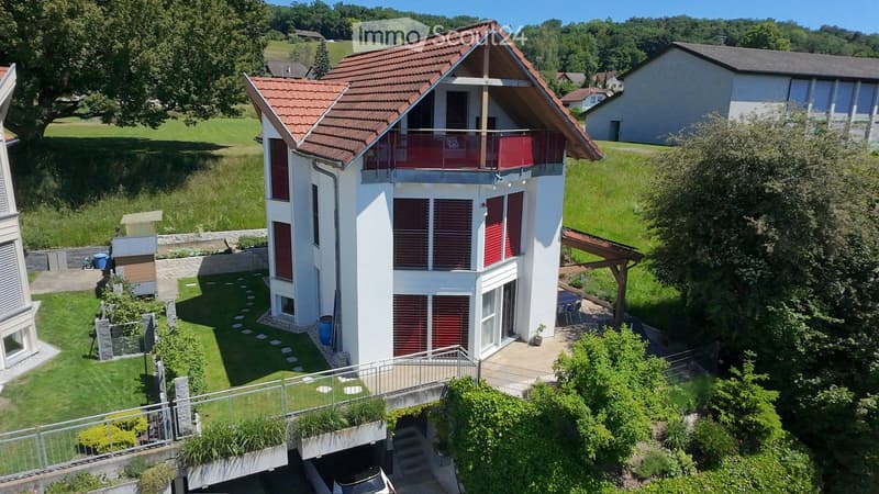 Modern two-story house with red-tiled roof, white exterior, and balcony overlooking a green landscape.