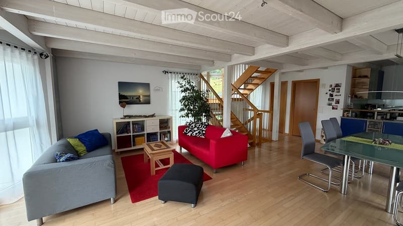 Open-plan living and dining area with wooden flooring, exposed wooden beams, a grey sofa, red sofa, coffee table, ottoman, dining table with chairs, bookshelf, and a staircase leading to an upper level.