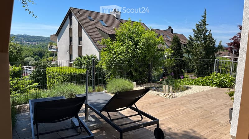 A wooden deck with two black lounge chairs, surrounded by a metal fence and lush greenery, with a modern house and scenic landscape in the background.