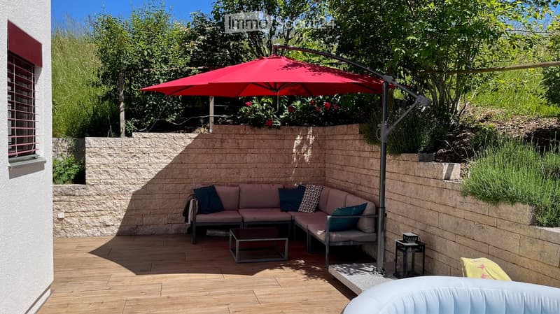 A modern outdoor seating area with a wooden deck, a grey sectional sofa, throw pillows, a coffee table, and a large red umbrella providing shade. The space is enclosed by a stone wall and features lush greenery and plants.