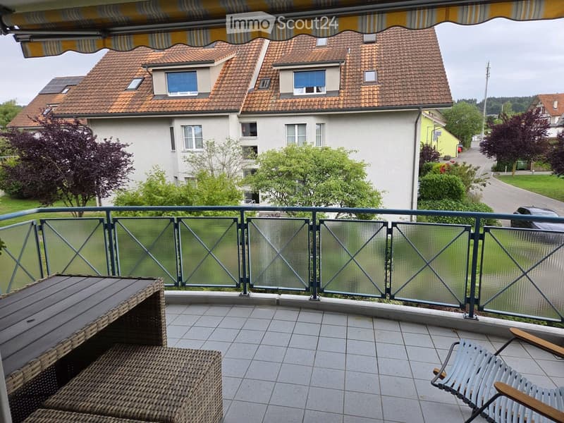Balcony with tiled floor, glass railing, and outdoor furniture including a wicker table and a folding chair.
