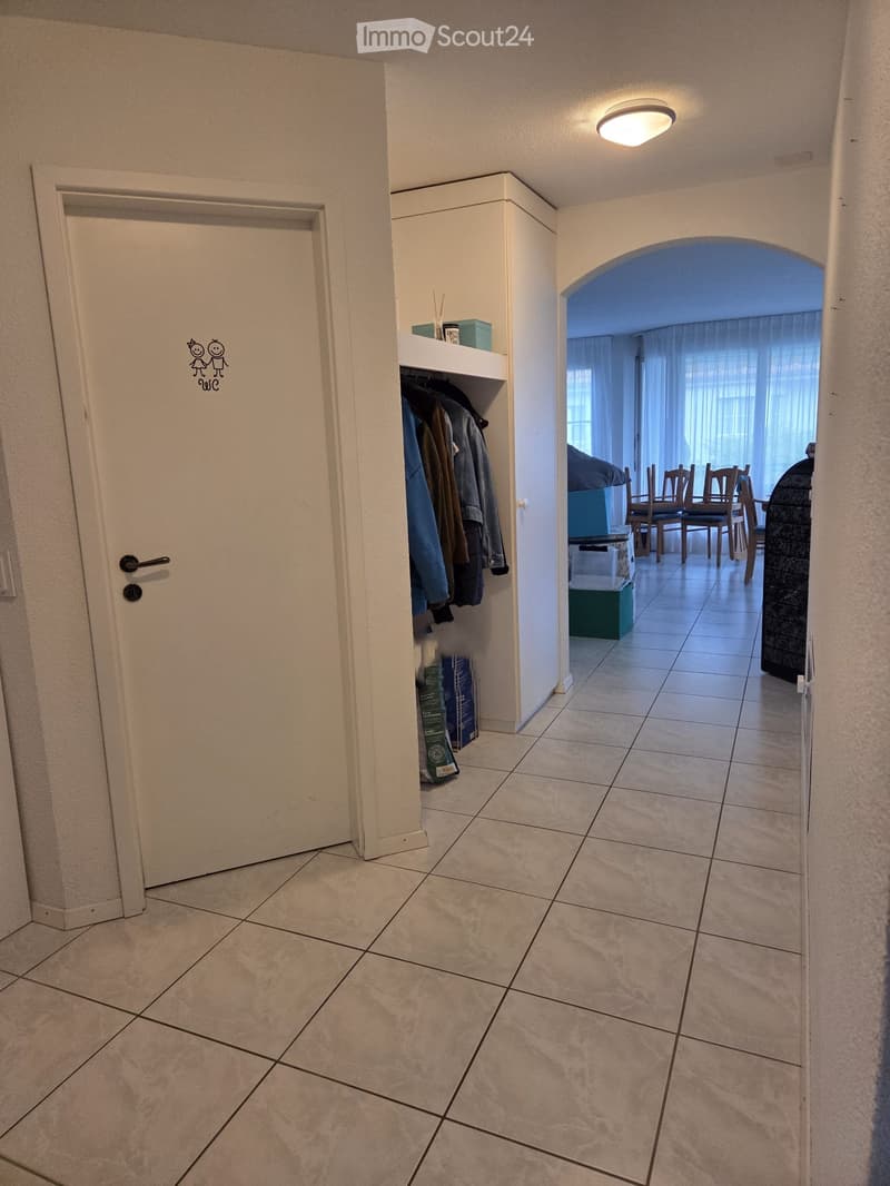 Hallway with tiled floor, white walls, a coat rack with jackets, and an open doorway leading to a dining area with chairs and a table.