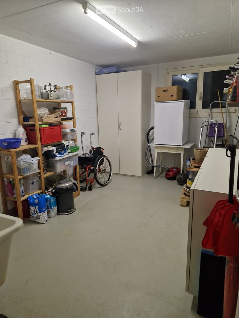 A small, functional room with a wooden shelf holding various items, a white refrigerator, a wheelchair, a cabinet, and a small table with a basket on it. The room has a concrete floor and white brick walls.
