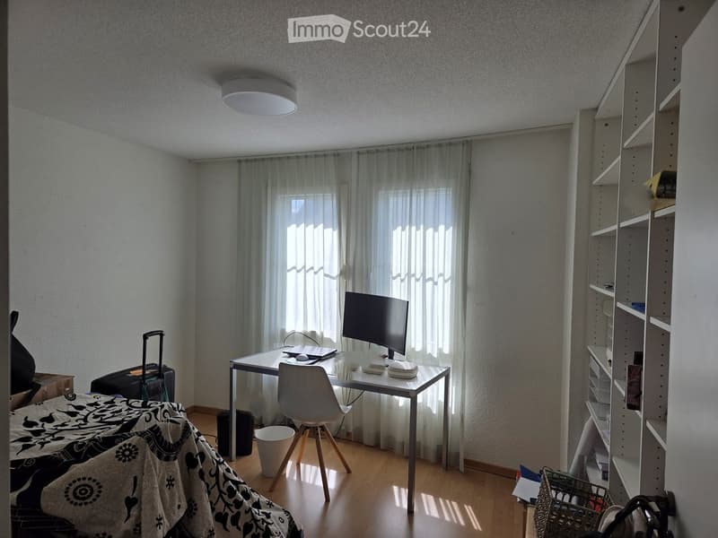 A furnished room with a bed, desk, chair, monitor, laptop, shelf, and curtains. The room has wooden flooring and a window with sheer curtains.