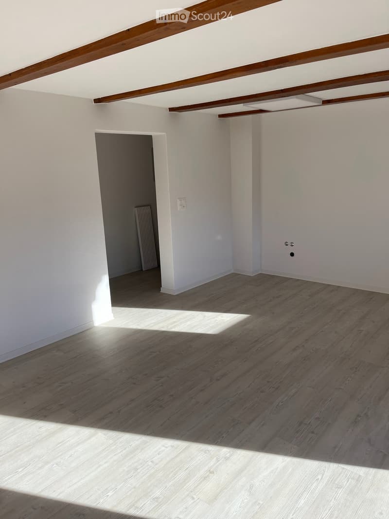 Empty room with light wooden flooring, white walls, and exposed wooden beams on the ceiling. A doorway leads to another room with a radiator visible.